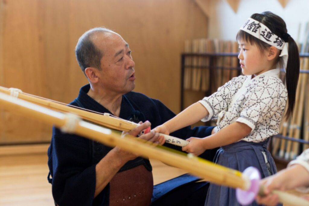 Tokyo Kyumeikan dojo Sensei padeda mergaitei taisyklingai laikyti shinai per vaikų kendo treniruotę.