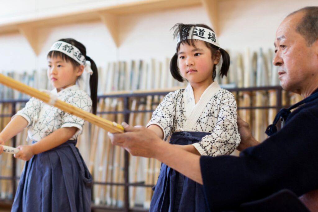Tokyo Kyumeikan instruktorius padeda dviems mergaitės atlikti kendo pratimą